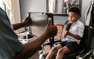 Young child sitting calmly in a barber chair while a stylist shows him his haircut in a mirror, creating a positive and supportive haircut experience for a child with sensory sensitivities.