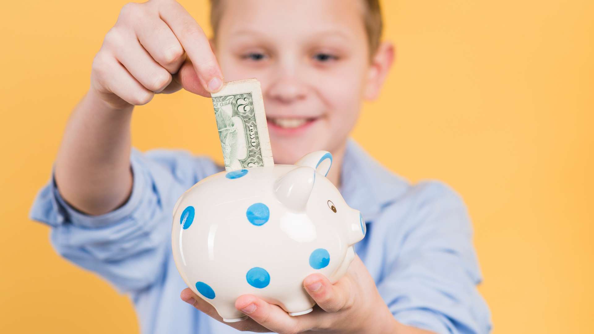 Smiling young boy proudly putting a dollar bill into a spotted piggy bank, practicing the money saving skills children with autism can build through early financial education