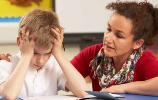 Frustrated young boy with autism holding his head during a classroom task while a compassionate teacher offers calm support and guidance