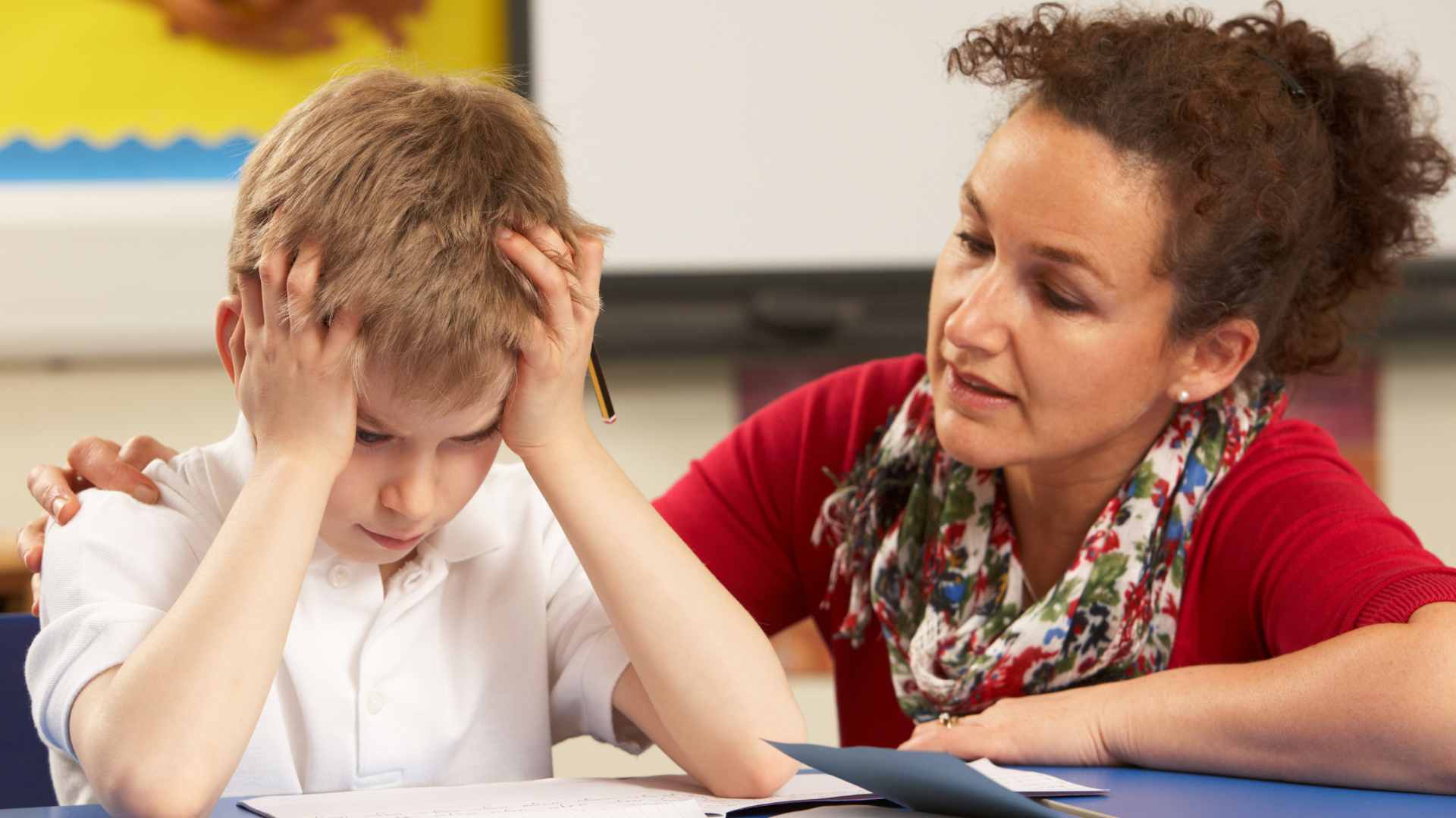 Frustrated young boy with autism holding his head during a classroom task while a compassionate teacher offers calm support and guidance